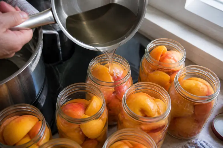 Hände gießen heißen Zuckersirup in ein sterilisiertes Glas mit Pfirsichhälften, während des Einmachprozesses in einer deutschen Küche.
