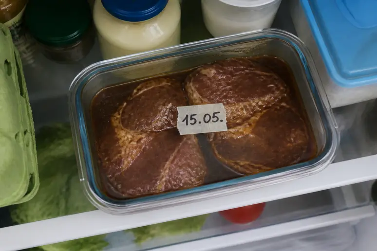 Ein gut belebter Kühlschrank mit einem Behälter, der mariniertes Fleisch und die Beize enthält, gut sichtbar.